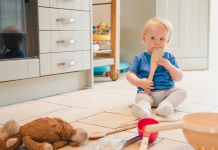 Toddler sitting on the kitchen floor playing with a wooden spoon, other kitchen items and a stuffed animal. Keep your baby entertained with treasure baskets.