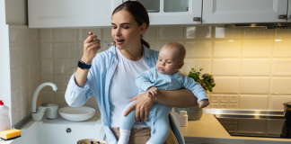 Photo of a mom eating over the sink while holding a baby. Postpartum nutrition isn't easy.