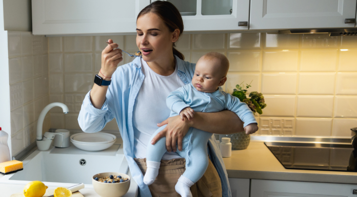 Photo of a mom eating over the sink while holding a baby. Postpartum nutrition isn't easy.