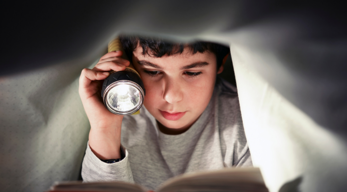 Young reader holding a flashlight and reading a story for book lovers under the covers.