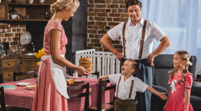A blonde woman presents a plate of pancakes to her husband and two children.