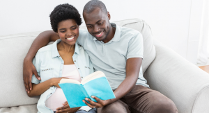 Two people sitting on a couch. One person has their arm around a pregnant person and they are reading a book together. Books to read when you're pregnant.
