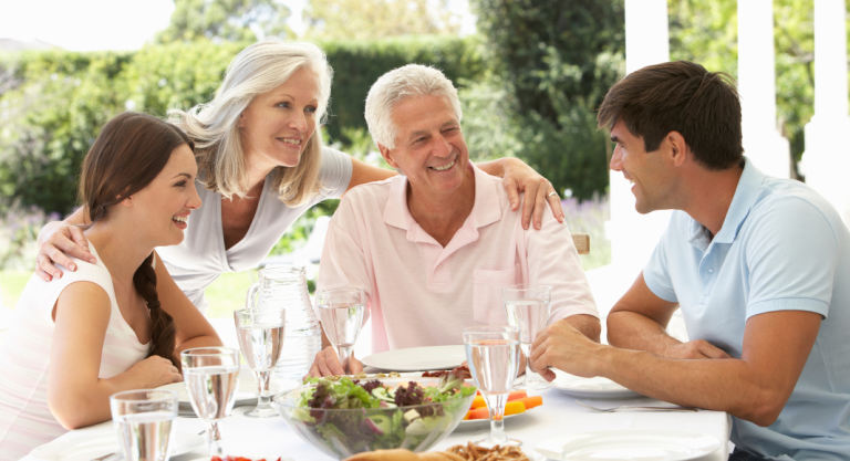 A family sitting around a table, daughter, mother, father and son. The children are young adults, the parents are older. They are close and smiling, they look happy to be together.